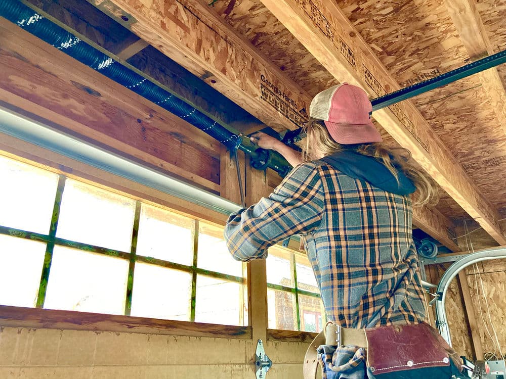 Person installing plumbing in a wooden structure with tools, wearing a plaid shirt and cap.